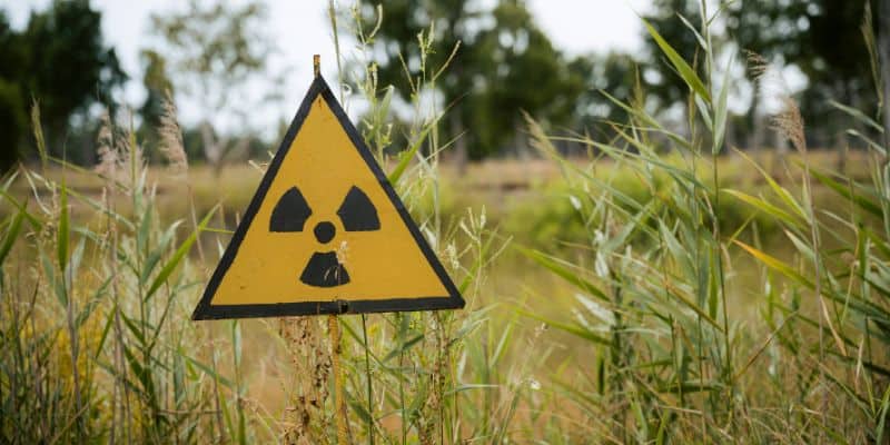 Auf dem Bild ist ein gelbes Warnschild mit dem Symbol für Radioaktivität zu sehen, das zwischen hohem Gras in einer verlassenen Landschaft steht.