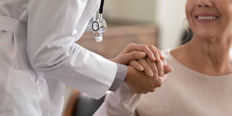 Ärztin schenkt Hoffnung. Nahaufnahme einer jungen Ärztin, die sich zu einer lächelnden älteren Patientin vorbeugt und ihre Hand in ihren Händen hält. Die Betreuerin im weißen Kittel unterstützt und ermutigt die ältere Frau.