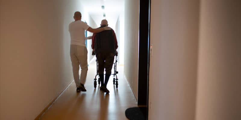 Gott im Alltag - Ein jüngerer Mann begleitet einen älteren, pflegebedürftigen Mann über den Flur
